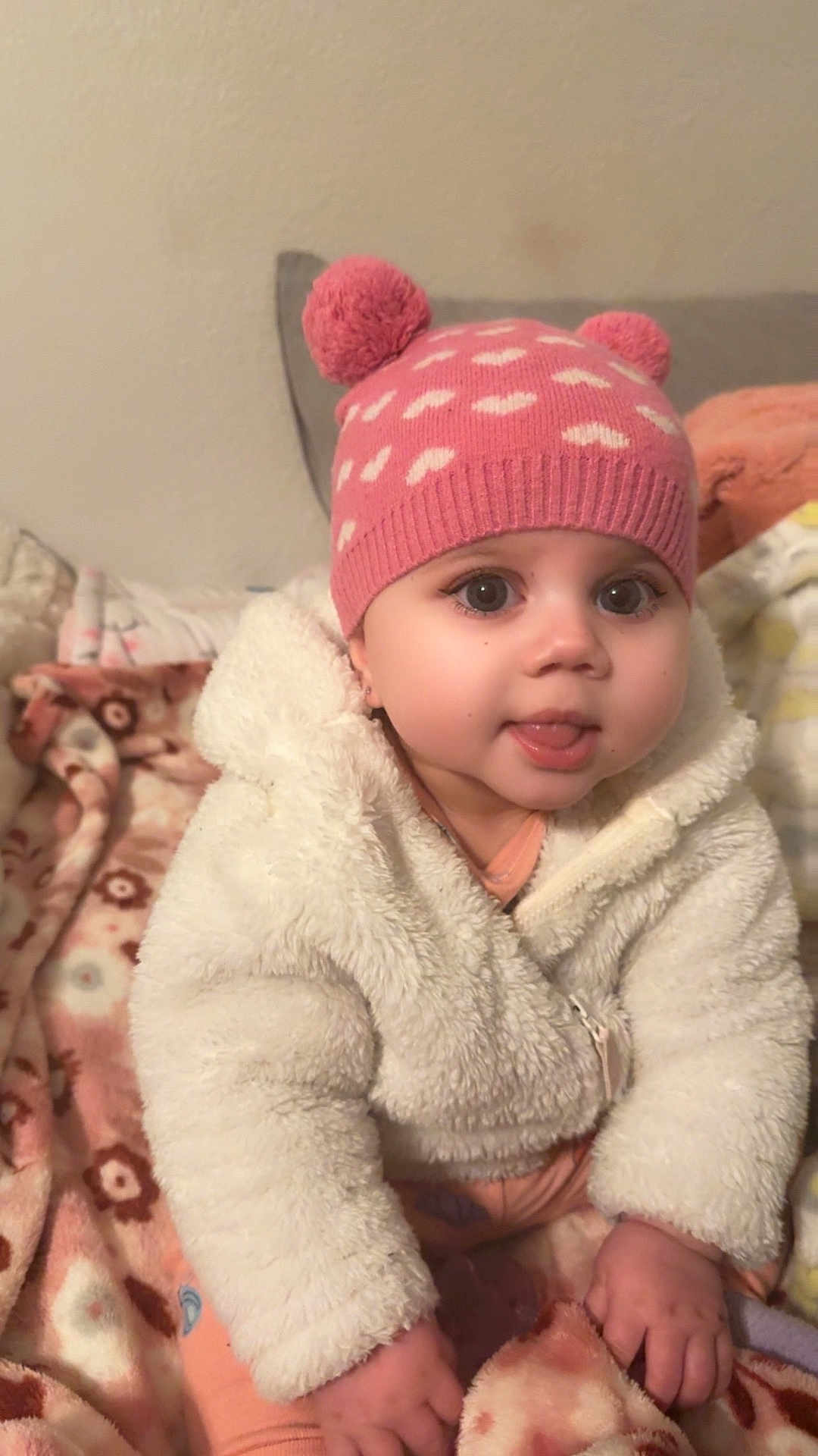 Gianna joined the competition — help win amazing prizes! baby, child, pink_hat, pom_poms, white_jacket, fluffy_clothing, blanket, patterned_blanket, indoor, cute, tongue_out, smiling, warm_clothing, seated, infant, face, hands, eyes, cozy, young_child