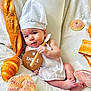 baby, infant, chef_hat, spatula, bread, pastry, croissant, baguette, doughnut, white_clothing, cute, smiling, lying_down, food, bakery, soft_fabric, indoors, portrait, three_months, props