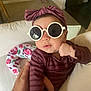 baby, child, sunglasses, headband, striped_clothing, hand, couch, floral_pillow, indoor, person, infant, face, fashion, accessory, comfortable, home, portrait, cute, holding, relaxed