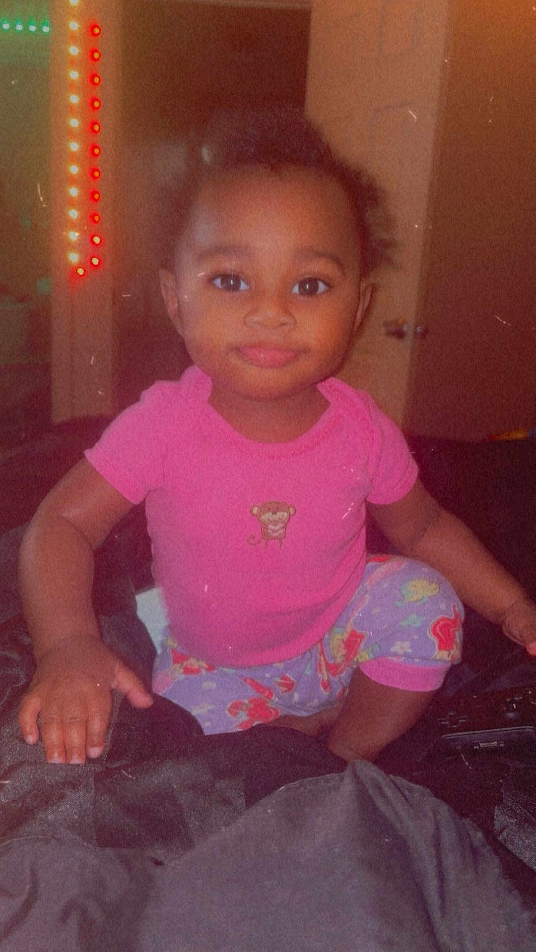 Tymaree Johnson is registered to the contest to win money with this photo: toddler, child, pink_shirt, pajamas, bed, indoor, smile, face, hand, room, colorful_lights, door, dark_background, cute, young_child, person, sitting, looking, cozy, casual_clothing