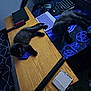 black_cat, kitten, desk, computer_keyboard, mouse, mousepad, hairbrush, notebook, bicycle_wheel, wooden_surface, purple_knitted_toy, bedspread, indoor, sleeping, lying_down, pet, technology, gaming_mouse, clutter, relaxation