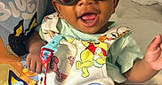 Michael joined the competition — help win amazing prizes! adult_hand, baby, bed, bedroom, cartoon_clothing, child, colorful, face, happy, held, indoor, infant, overalls, pacifier_clip, photography, portrait, sitting, smiling, sunglasses, toy