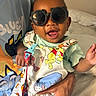adult_hand, baby, bed, bedroom, cartoon_clothing, child, colorful, face, happy, held, indoor, infant, overalls, pacifier_clip, photography, portrait, sitting, smiling, sunglasses, toy