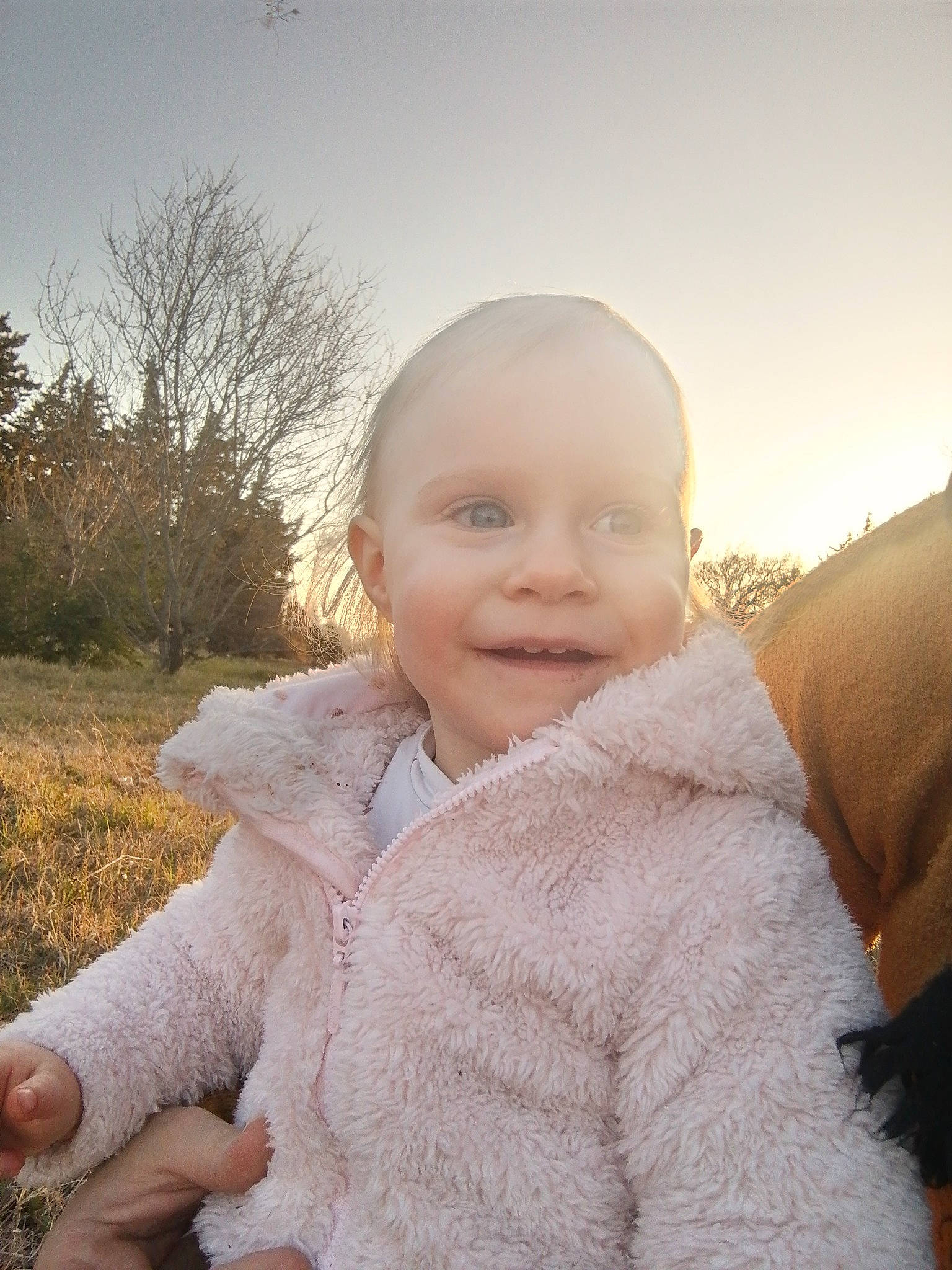 Lucie a rejoint le concours — aidez-le/la à gagner de superbes lots ! baby, baby_toddler_clothing, finger, fun, gesture, grass, happy, iris, joy, outerwear, people_in_nature, person, plant, sitting, sky, sleeve, smile, toddler, tree, winter