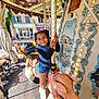 amusement_ride, black_sleeves, blue_clothing, braided_hair, building, carousel, child, daylight, decorative_lights, fun, outdoor, pastoral_painting, pig, playful, smiling, urban_background, vintage, white_shoes, window, wooden_floor