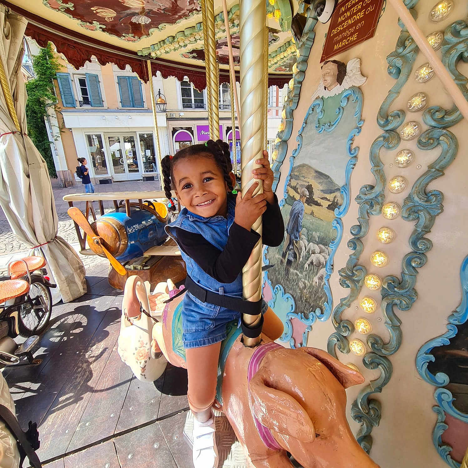 Melyne Marie participe au concours pour gagner de l'argent avec cette photo : child, smiling, carousel, pig, amusement_ride, outdoor, daylight, braided_hair, blue_clothing, black_sleeves, white_shoes, vintage, decorative_lights, pastoral_painting, urban_background, window, building, wooden_floor, fun, playful