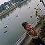 child, girl, sunglasses, river, water, ducks, guardrail, plants, grass, buildings, cars, outdoor, nature, casual_clothing, playful, summer, footwear, curly_hair, urban, daytime