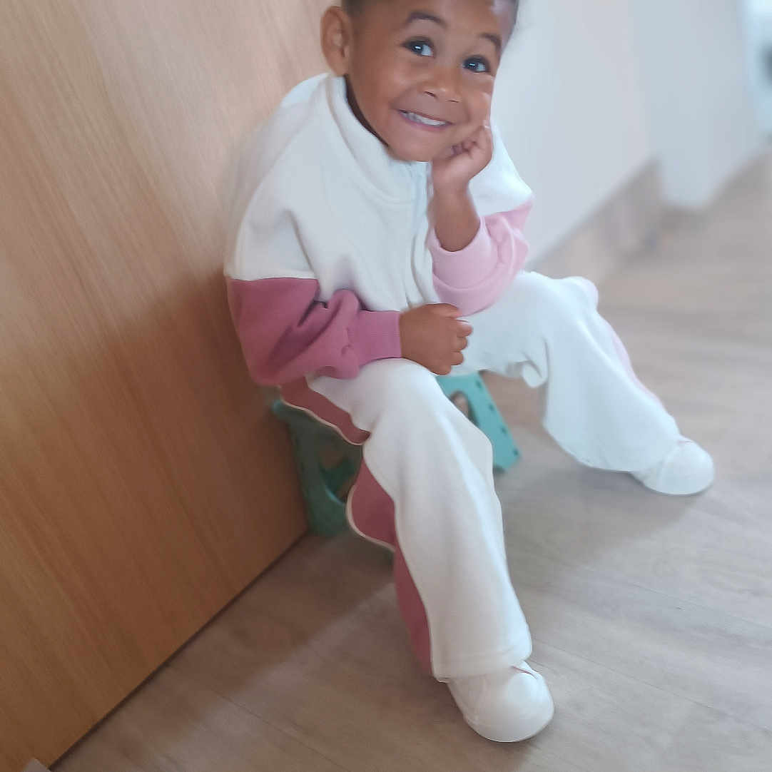 Melyne Marie a rejoint le concours — aidez-le/la à gagner de superbes lots ! blurred_background, casual_clothing, child, curly_hair, floor, flooring, happy, home, indoor, light, person, playful, portrait, relaxed, seated, smiling, stool, white_shoes, wooden_door, young_child