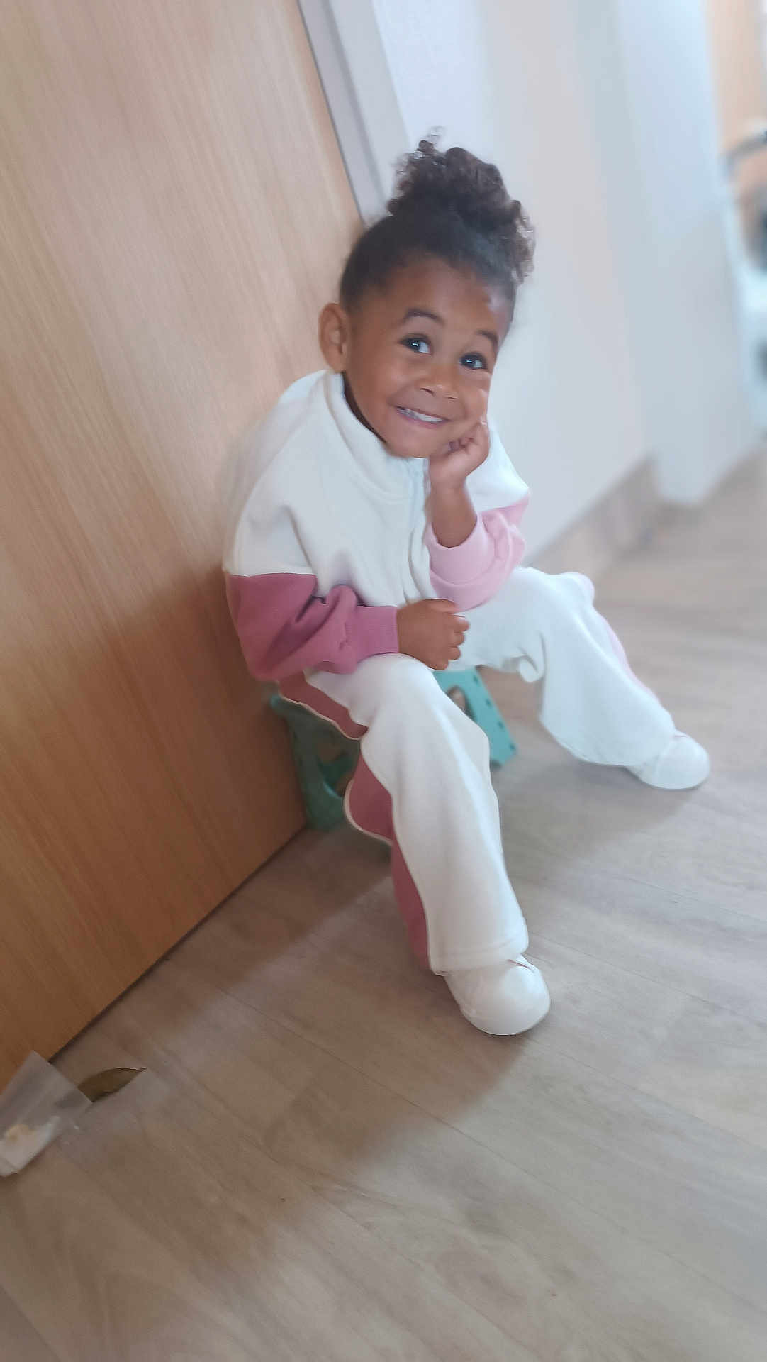 Melyne Marie a rejoint le concours — aidez-le/la à gagner de superbes lots ! blurred_background, casual_clothing, child, curly_hair, floor, flooring, happy, home, indoor, light, person, playful, portrait, relaxed, seated, smiling, stool, white_shoes, wooden_door, young_child
