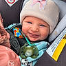 baby, smiling, car_seat, pacifier, hat, pink_bow, clothing, infant, child, safety_harness, seat_belt, cute, cozy, warm_clothing, indoor, portrait, happy, face, person, closeup