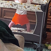 Penelope is registered to the contest to win money with this photo: baby, playpen, orange_shorts, living_room, television, text_overlay, toys, rug, wooden_cabinet, decor, child, furniture, floor, blanket, cushion, standing, reaching, indoor, colorful, screen