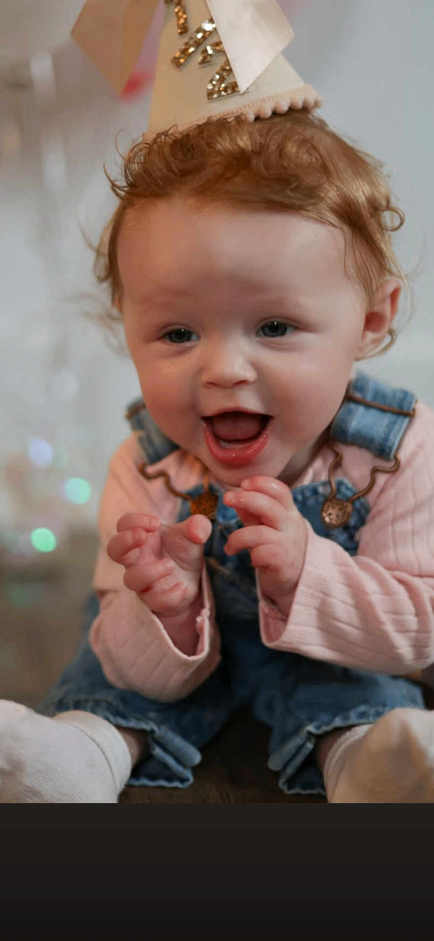 Aspen is registered to the contest to win money with this photo: baby, toddler, child, party_hat, birthday_hat, overalls, denim, pink_shirt, smiling, laughing, close_up, portrait, curly_hair, blue_eyes, hands, socks, indoor, soft_lighting, bokeh, festive