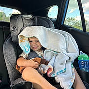 Eloy is registered to the contest to win money with this photo: child, toddler, smiling, car, car_seat, blanket, stuffed_animal, toy, water_bottle, road_trip, passenger, indoors, vehicle, portrait, sitting, casual, daylight, window, happy, family_travel