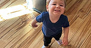 Eloy is registered to the contest to win money with this photo: child, toddler, smiling, portrait, standing, indoor, hardwood_floor, wooden_floor, cabinet, furniture, sunlight, natural_light, blue_shirt, black_pants, socks, reebok, blond_hair, face, home, happy