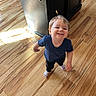 child, toddler, smiling, portrait, standing, indoor, hardwood_floor, wooden_floor, cabinet, furniture, sunlight, natural_light, blue_shirt, black_pants, socks, reebok, blond_hair, face, home, happy