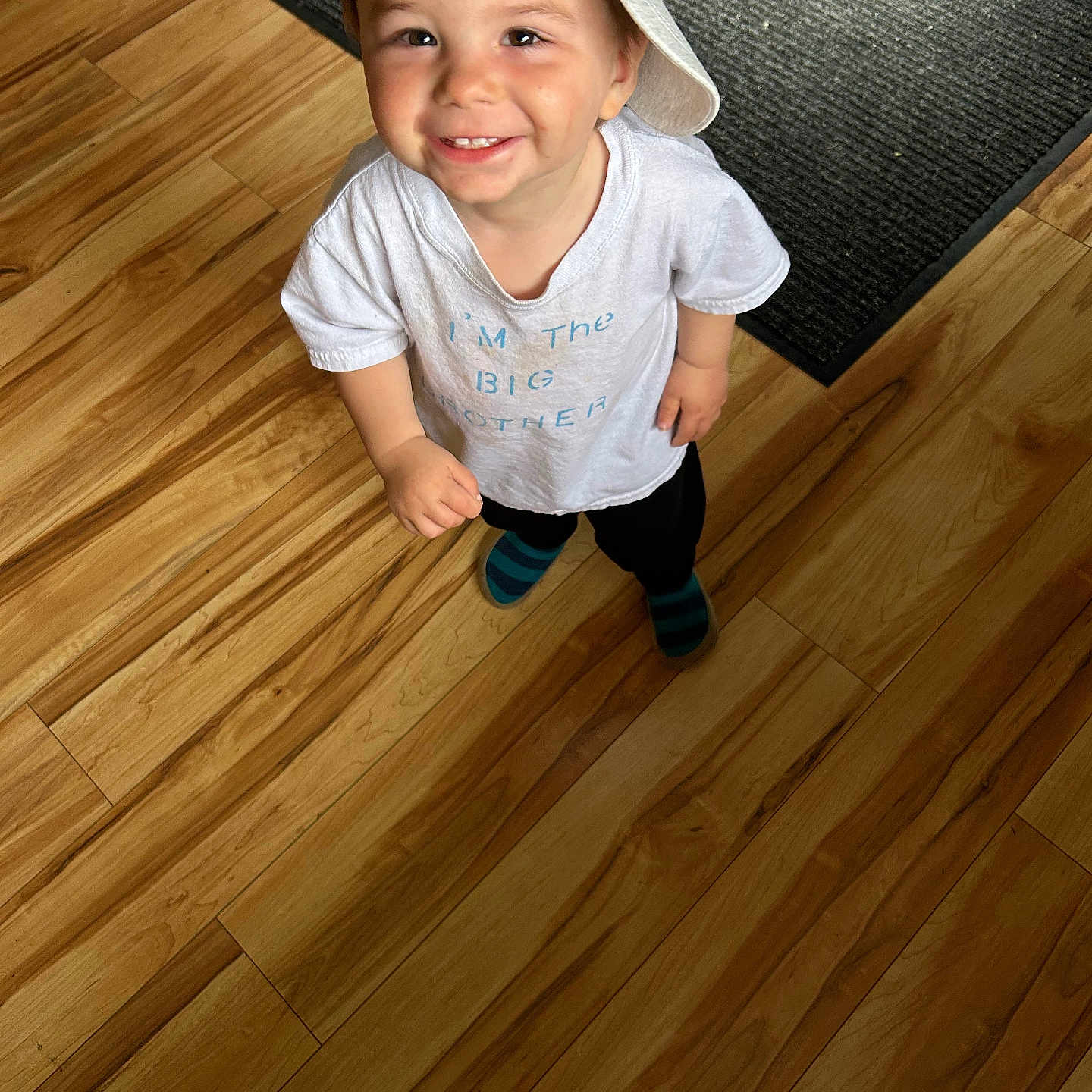Eloy is registered to the contest to win money with this photo: child, toddler, smiling, happy, portrait, indoor, wooden_floor, rug, hat, patterned_hat, white_shirt, black_pants, striped_socks, standing, looking_at_camera, brown_hair, face, home, flooring, cute