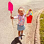 child, girl, smiling, outdoor, pavement, shovel, bucket, shadow, sunlight, skirt, long_sleeve, floral, play, toy, happy, casual_clothing, footwear, greenery, sidewalk, daytime