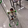 beverages, bottles, child, floor, food, green_cart, gripping, grocery_store, indoor, juice, portrait, retail, seated, shelves, shopping, shopping_cart, small_child, store, supermarket, toddler