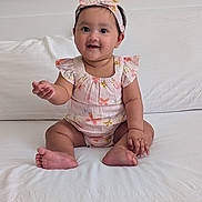 Isra is registered to the contest to win money with this photo: baby, child, smiling, sitting, headband, romper, butterfly_pattern, indoor, bed, white_bedspread, cute, happy, person, infant, young_child, clothing, skin, hand, foot, portrait