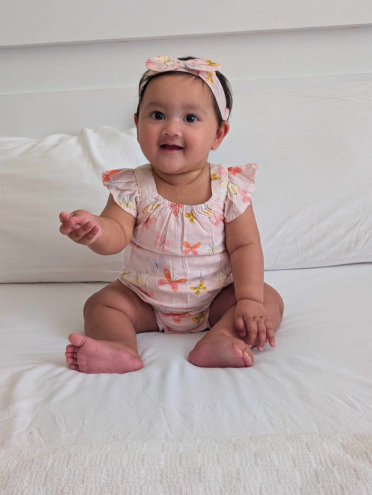 Isra is registered to the contest to win money with this photo: baby, child, smiling, sitting, headband, romper, butterfly_pattern, indoor, bed, white_bedspread, cute, happy, person, infant, young_child, clothing, skin, hand, foot, portrait