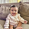 baby, child, smiling, happy, portrait, sitting, couch, sofa, pillow, living_room, indoors, home, infant, girl, bow, sweater, pink_clothing, barefoot, curly_hair, natural_light