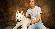 Rex participe au concours pour gagner de l'argent avec cette photo : backdrop, blue_eyes, casual_clothing, companion, dog, glasses, happy, indoor, leash, man, person, pet, photography, portrait, seated, smiling, studio, tongue, white_dog, wooden_bench