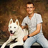 backdrop, blue_eyes, casual_clothing, companion, dog, glasses, happy, indoor, leash, man, person, pet, photography, portrait, seated, smiling, studio, tongue, white_dog, wooden_bench