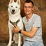 animal_photography, backdrop, casual_clothing, companion, dog, glasses, harness, human, indoors, leash, man, pet_owner, portrait, sitting, smiling, standing_dog, studio, watch, white_dog, wood_floor