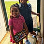 child, girl, smiling, braided_hair, beads, tablet, cartoon, doorway, sunlight, indoor, dress, sneakers, carpet, happy, family, technology, play, portrait, sibling, casual