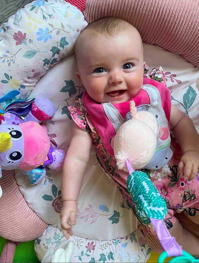 Katie joined the competition — help win amazing prizes! baby, child, smiling, plush_toy, cushion, floral_pattern, pink_clothing, bib, indoor, playtime, cute, happy, infant, person, soft_toys, colorful, lying_down, head, hand, face