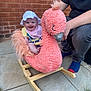baby, child, rocking_toy, flamingo, pink, hat, bib, smiling, outdoor, patio, brick_wall, adult, jeans, blue_socks, grass, toy, happy, playful, person, footwear
