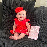 Katie is registered to the contest to win money with this photo: baby, infant, red_dress, headband, flower, couch, pillow, milestone_card, smiling, sitting, indoors, child, cute, happy, portrait, person, feet, skin, clothing, home