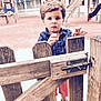 child, playground, wooden_gate, latch, fence, outdoor, person, shoe, shorts, shirt, lock, hand, face, expression, play_area, daylight, young, curly_hair, standing, background