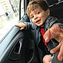 child, boy, car_interior, backseat, window, smile, hand, seatbelt, jacket, shorts, playful, portrait, vehicle, urban, rainy, street, people, door_handle, window_glass, casual_clothing