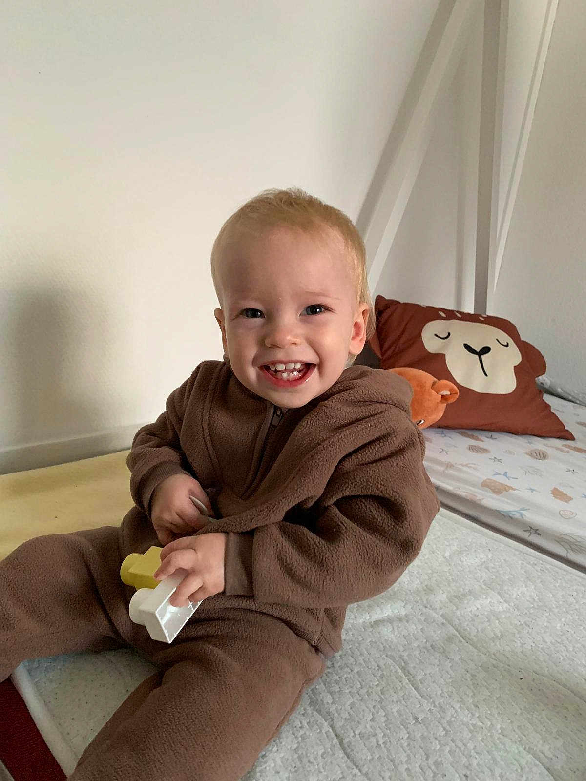 Aaron participe au concours pour gagner de l'argent avec cette photo : toddler, child, smile, toy, bed, pillow, brown_clothing, indoor, happy, fleece, face, person, cute, playful, home, furniture, baby, sitting, room, cozy