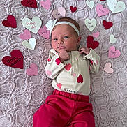 Birdie is registered to the contest to win money with this photo: baby, newborn, infant, headband, paper_hearts, pink_blanket, valentine, onesie, heart_pattern, red_pants, lying_down, portrait, cute, small_hands, soft_texture, nursery, decor, celebration, closeup_face, fabric