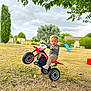 toddler, child, toy_motorcycle, wheelie, outdoor, grass, tree, sky, cloudy, house, chair, play, fun, blonde_hair, young_child, rural, nature, greenery, daytime, casual_clothing