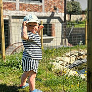 Gabriel a rejoint le concours — aidez-le/la à gagner de superbes lots ! toddler, child, boy, striped_clothing, hat, cap, outdoor, garden, grass, flowers, sunlight, shoes, fence, brick_house, daytime, nature, portrait, standing, smiling, playful