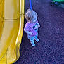 toddler, child, playground, slide, yellow_slide, pink_shirt, patterned_pants, curly_hair, sneakers, outdoor, play_area, rubber_surface, blue_pole, green_slide, standing, looking_sideways, daylight, casual_clothing, young_child, fun