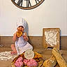 Milann participe au concours pour gagner de l'argent avec cette photo : baby, child, chef_hat, apron, croissant, cookies, heart_shaped, pink_frosting, bread, baguette, wooden_board, flour, whisk, clock, kitchen_counter, sitting, eating, smiling, indoor, baking