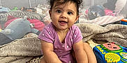 Maylani joined the competition — help win amazing prizes! baby, child, smiling, curly_hair, purple_onesie, sitting, toys, blanket, quilt, indoor, play_area, cute, hands, feet, face, happy, infant, colorful, playtime, home