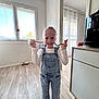 braids, cabinet, child, curtains, flooring, girl, happy, home, indoor, natural_light, overalls, person, playful, room, smile, socks, standing, white_shirt, window, young