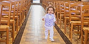 Elyo a rejoint le concours — aidez-le/la à gagner de superbes lots ! aisle, background, bowtie, chairs, child, church, curly_hair, formal_wear, happy, indoor, light, person, playful, smiling, tile_floor, toddler, vest, walking, wooden_chairs, young_child