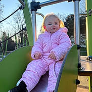 Leïa a rejoint le concours — aidez-le/la à gagner de superbes lots ! backlit, boots, child, cold_weather, happy, metal_structure, outdoor, park, pink_jacket, play_equipment, playground, railings, sitting, slide, smile, sunlight, toddler, toy_car, trees, winter_coat