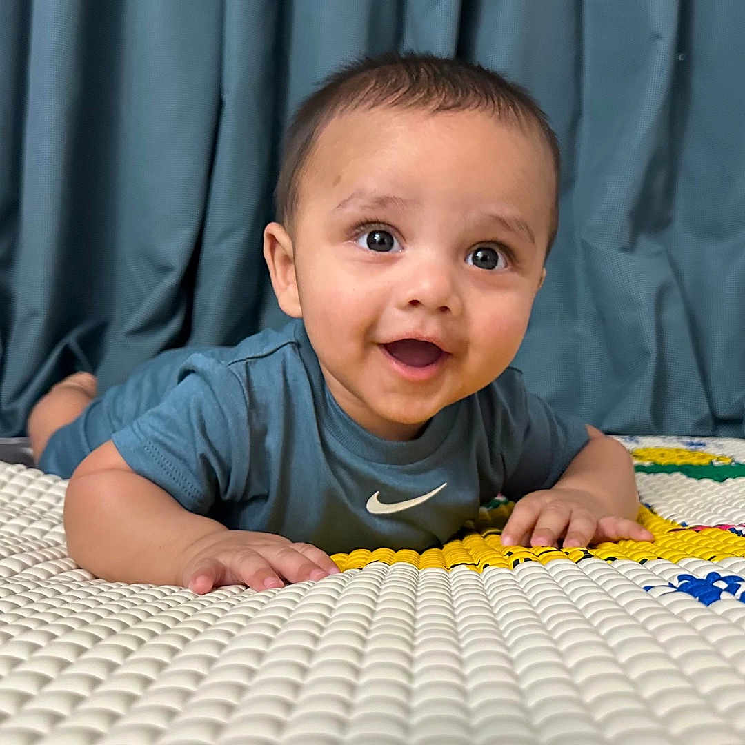 Nived is registered to the contest to win money with this photo: baby, child, toddler, smiling, indoor, play_mat, curtains, blue, happy, person, face, clothing, short_hair, skin, hands, lying_down, expression, cute, young, portrait