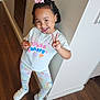 child, girl, peace_sign, smile, bow, snack, leggings, tshirt, indoor, floor, wooden_floor, wall, hallway, happy, footwear, sneakers, curly_hair, earrings, pose, casual_clothing