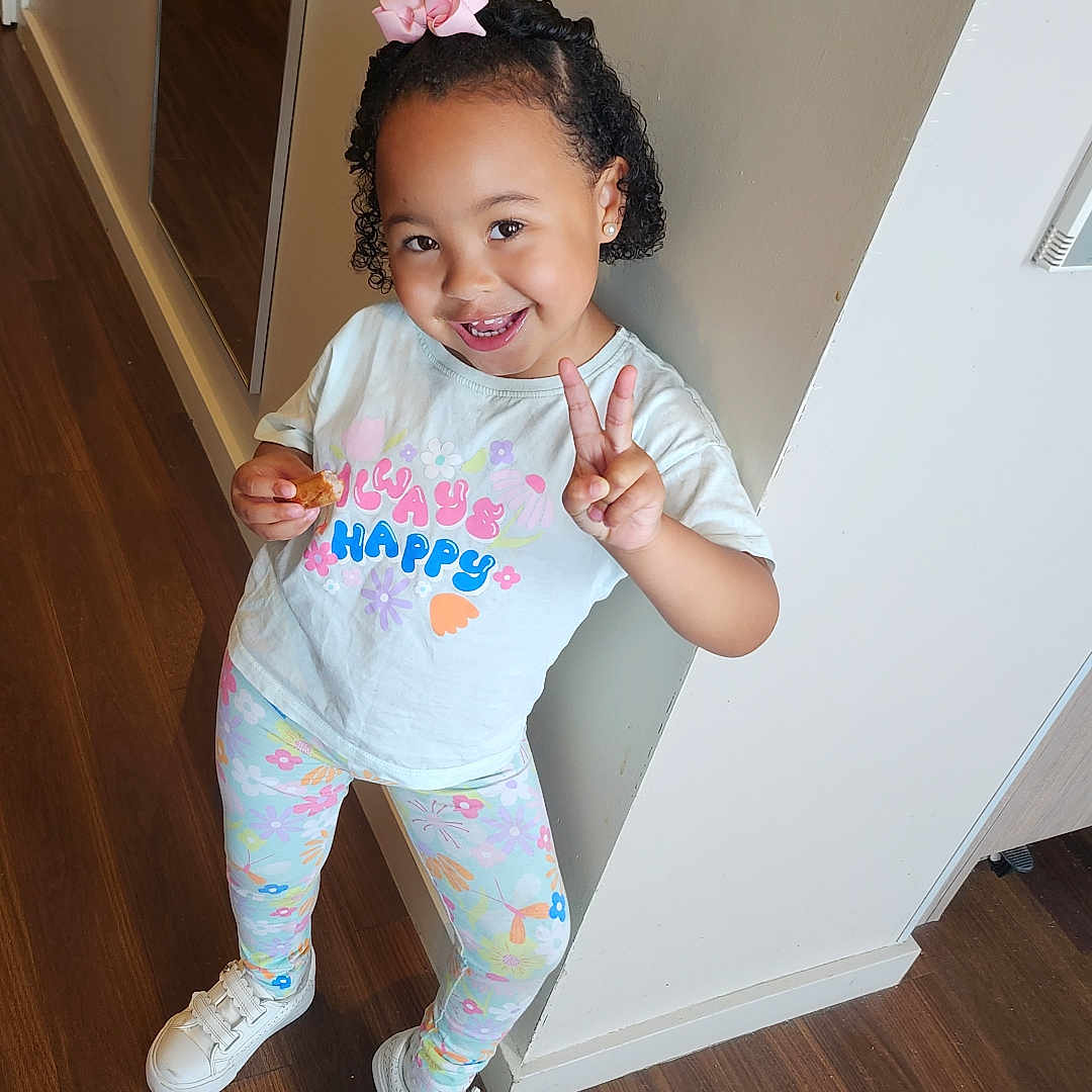 Renayah-Marie is registered to the contest to win money with this photo: bow, casual_clothing, child, curly_hair, earrings, floor, footwear, girl, hallway, happy, indoor, leggings, peace_sign, pose, smile, snack, sneakers, tshirt, wall, wooden_floor