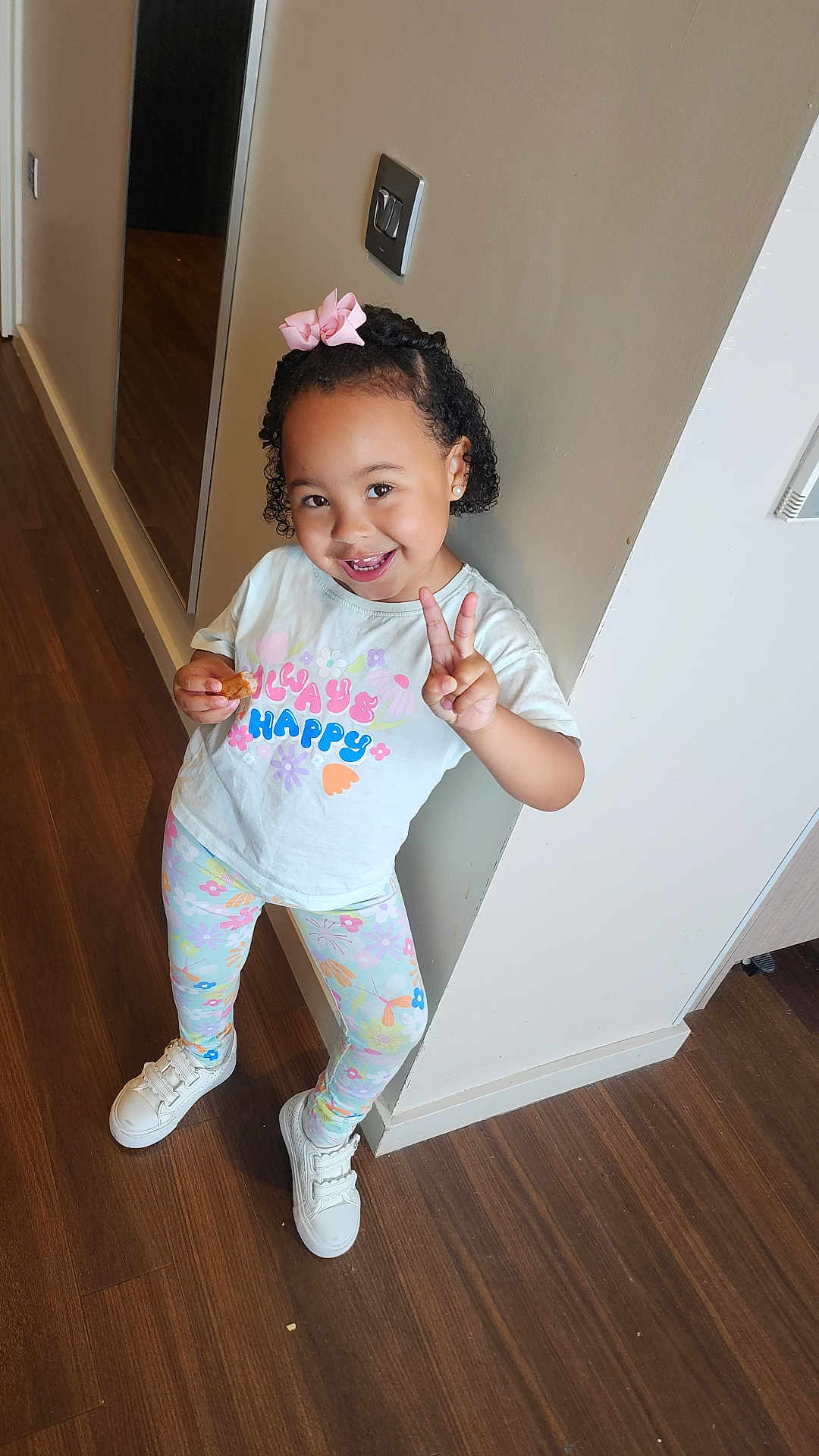 Renayah-Marie is registered to the contest to win money with this photo: child, girl, peace_sign, smile, bow, snack, leggings, tshirt, indoor, floor, wooden_floor, wall, hallway, happy, footwear, sneakers, curly_hair, earrings, pose, casual_clothing
