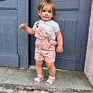 Julia a rejoint le concours — aidez-le/la à gagner de superbes lots ! toddler, child, standing, outdoor, door, blue_door, sandals, pink_overall, daisy_pattern, shorts, white_tshirt, curly_hair, sidewalk, concrete, expression, person, young_child, summer_clothing, casual, portrait