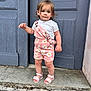 toddler, child, standing, outdoor, door, blue_door, sandals, pink_overall, daisy_pattern, shorts, white_tshirt, curly_hair, sidewalk, concrete, expression, person, young_child, summer_clothing, casual, portrait
